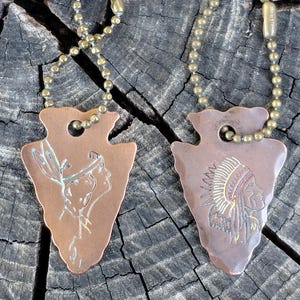 May include: Two copper-colored arrowhead pendants with brass ball chains. One pendant features an etched Native American profile with a feathered headdress. The other pendant has a profile of a Native American with a headdress. The pendants are displayed on a weathered wooden surface.