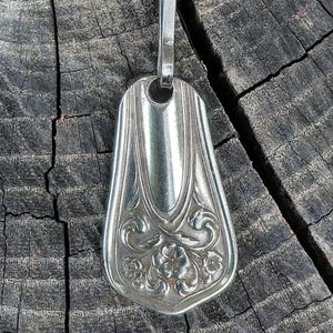 May include: A silver pendant crafted from a repurposed spoon, featuring a floral design on the spoon's bowl. The pendant has a loop for a chain and is displayed against a rustic wooden background.