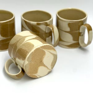 May include: Four ceramic mugs with a unique marbled design. The mugs feature a tan and cream swirl pattern, with a cylindrical shape and a curved handle. The mugs are set against a white background, showcasing their earthy tones and handmade quality.
