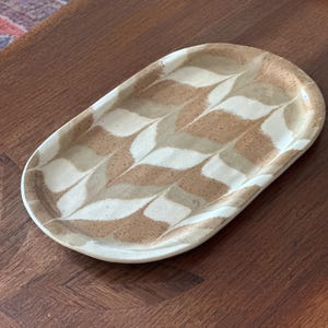 Op de afbeelding: Ovale keramische schaal met een bladerenpatroon in bruin, beige en wit. De schaal heeft een licht verhoogde rand en een gespikkelde textuur. Het ontwerp is een herhalend patroon van gestileerde bladeren. Geschikt voor serveren of presenteren.