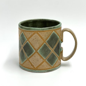 Puede incluir: Taza de cerámica con un patrón de diamantes en beige, verde y marrón. La taza tiene un asa redondeada y un interior verde. El exterior presenta un patrón repetido de diamantes verdes delineados en marrón sobre un fondo beige.