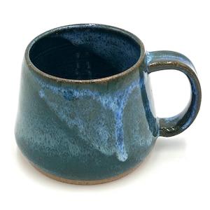 Puede incluir: Una taza de cerámica hecha a mano con una forma única y orgánica. La taza presenta un esmalte verde azulado oscuro con vetas azules y blancas, y una base y asa marrones. El interior es azul oscuro.