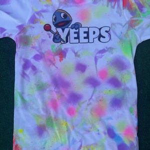 May include: White t-shirt with a colorful spray-paint design in pink, purple, green, and orange. The shirt features a cartoon character holding a lollipop and the word "YEEPS" in bold, white letters.