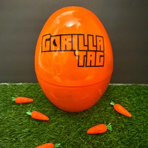 以下が含まれることがあります： 「GORILLA TAG」と印刷された大きなオレンジ色のプラスチック製の卵。卵は緑色の芝生の上に置かれ、その周りに小さなオレンジ色のニンジンが6本あります。