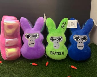 May include: Four plush bunny-shaped pillows with different colors and personalized names. The pillows are pink, purple, green, and blue. The green pillow has the name "Brayden" and the year "2023" on it.