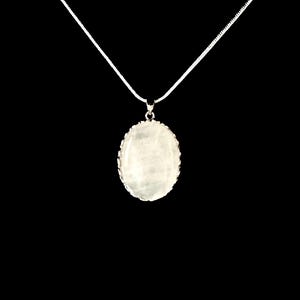 May include: A silver necklace with a white, oval-shaped pendant. The pendant has a textured surface and a decorative silver border. The chain is thin and delicate, and the background is black.