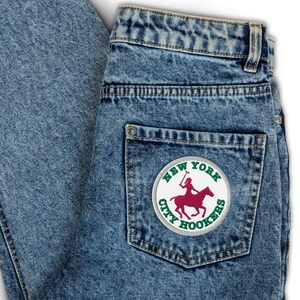 Puede incluir: Vaqueros de mezclilla azul claro con un parche en el bolsillo trasero. El parche circular tiene las palabras "NEW YORK CITY HOOKERS" en verde. Una figura granate sobre un caballo está en el centro.