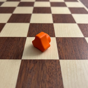 May include: An orange plastic game piece shaped like a rounded triangle sits on a wooden chessboard.