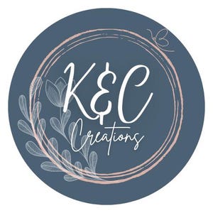 May include: A blue circle with a pink and white double circle frame. The text "K&C Creations" is in white inside the frame. There are white floral designs around the frame.