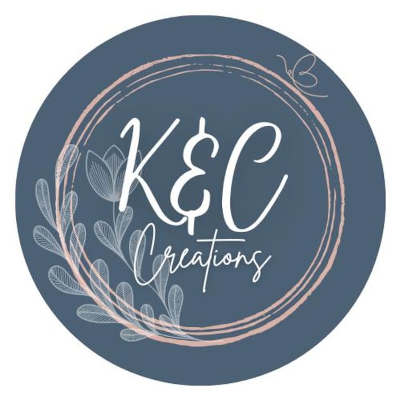 May include: A blue circle with a pink and white double circle frame. The text "K&C Creations" is in white inside the frame. There are white floral designs around the frame.
