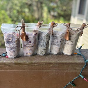 May include: Five clear pouches filled with bath salts, each sealed with a wooden scoop and twine. The pouches contain different colored salts and dried botanicals. A string of colorful lights is in the foreground.