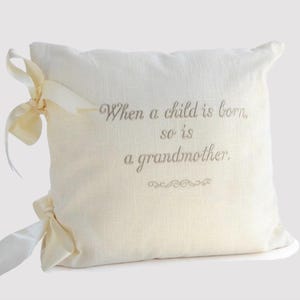May include: Cream-colored decorative pillow with the embroidered text: "When a child is born, so is a grandmother." The pillow has cream-colored ribbons tied on the sides. A thoughtful gift for a new grandmother.