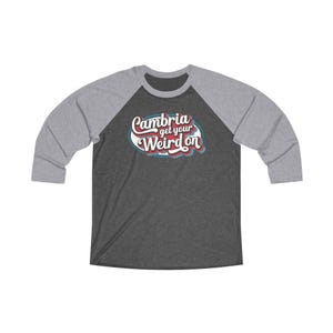 Puede incluir: Una camiseta de béisbol de color gris jaspeado y blanco con un gráfico de estilo retro que dice "Cambria get your weird on".