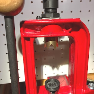 May include: A red metal reloading press featuring a black handle with a wooden knob, a black adjustment knob at the top, and a brass-colored pointed die component. The press has a sturdy frame with silver-colored bolts and a black circular base insert.