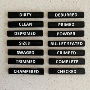 May include: A set of fourteen rectangular black labels with white text arranged in two columns. The labels feature the words: DIRTY, CLEAN, DEPRIMED, SIZED, SWAGED, TRIMMED, CHAMFERED, DEBURRED, PRIMED, POWDER, BULLET SEATED, CRIMPED, COMPLETE, and CHECKED.