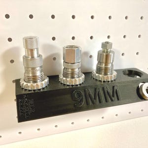 May include: A black 3D-printed storage block labelled 9MM with three silver metal reloading dies inserted into the top. The block features a textured side surface and a small circular cutout on the right side holding a single metal washer.