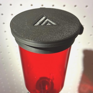 May include: A translucent red cylindrical container topped with a circular black lid. The lid features a textured surface and a recessed triangular logo design. The container has a smooth, glossy finish and is positioned vertically in the frame.