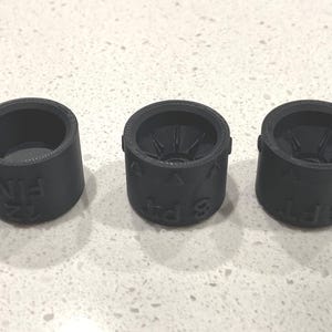 May include: Three black cylindrical objects with text and markings. The objects appear to be 3D printed. The text on the objects includes the numbers 6, 8, and 12, along with the letters "FT" and "FIN".