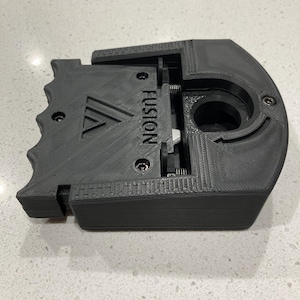 May include: A dark gray, 3D-printed device with the word "FUSION" and a "V" logo. The device has a complex mechanical design with multiple parts, including springs and a circular opening. The device is designed to fit a specific purpose.