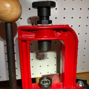 May include: A red reloading press with a wooden handle and black components. The press features a hexagonal black top and various metal parts, including bolts and a die holder. The background is a white pegboard.