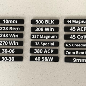 May include: A collection of eighteen rectangular black labels with white text displaying various ammunition calibers including 10mm, 223 Rem, 243 Win, 270 Win, 30-06, 30-30, 300 BLK, 308 Win, 357 Magnum, 38 Special, 380 ACP, 40 S&W, 44 Magnum, 45 ACP, 45 Colt, 6.5 Creedmoor, 7mm Rem Mag, and 9mm.
