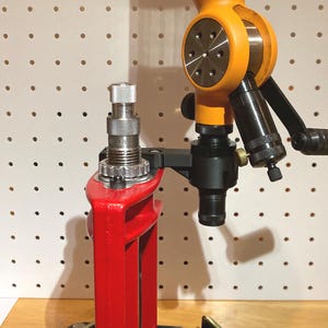 May include: A reloading press with a red frame and a silver die. An orange powder measure is attached, with a black handle and a black powder drop tube. The setup is on a wooden surface, against a white pegboard.