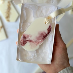 May include: An oval-shaped, cream-colored decorative item with a maroon, marbled design, presented in a white gift box with a cream-colored ribbon. The item is nestled in white tissue paper, suggesting a gift or decorative piece.