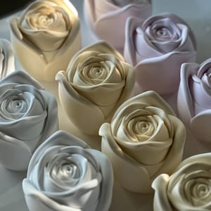 Puede incluir: Una colección de esculturas de rosas de cerámica en varios colores pastel, incluyendo crema, lavanda y azul pálido. Las rosas están intrincadamente detalladas, con cada pétalo cuidadosamente esculpido para parecerse a una flor real.