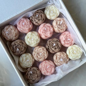 May include: A square white gift box containing sixteen flower-shaped candles arranged in a grid. The candles are molded to resemble peonies in various shades of cream, soft pink, and muted brown, each featuring a small white wick at the center.