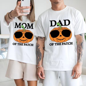 May include: Two white short-sleeved t-shirts featuring a graphic of an orange pumpkin wearing black sunglasses. The left shirt has the text MOM OF THE PATCH, and the right shirt has the text DAD OF THE PATCH printed in black above and below the pumpkin.