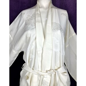 May include: Ivory-colored kimono-style robe with a floral pattern. The robe features long sleeves, a wrap-around design, and a matching belt. The fabric has a subtle sheen, and the floral design is visible throughout the garment.