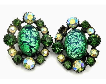 May include: A pair of green and clear glass earrings with a black metal setting. The earrings feature a large oval green glass stone surrounded by smaller clear and green stones.