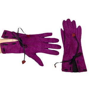 May include: A pair of magenta suede gloves. The gloves have black leather accents and a black tie detail at the wrist. A small, heart-shaped red leather accent is attached to the tie. The gloves are designed for cold weather.