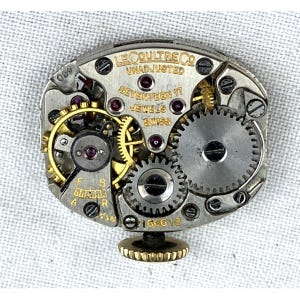 Op de afbeelding: Close-up van een uurwerk met ingewikkelde tandwielen, juwelen en een goudkleurige kroon. De tekst "LE COULTRE CO" en "SEVENTEEN 17 JEWELS SWISS" zijn zichtbaar. Het horlogemechanisme is zilver en goud.