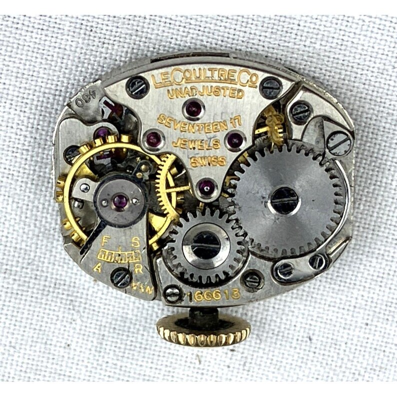 Op de afbeelding: Close-up van een uurwerk met ingewikkelde tandwielen, juwelen en een goudkleurige kroon. De tekst "LE COULTRE CO" en "SEVENTEEN 17 JEWELS SWISS" zijn zichtbaar. Het horlogemechanisme is zilver en goud.