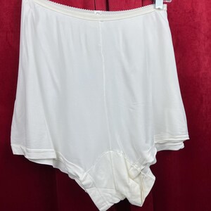 May include: Off-white vintage slip shorts. These high-waisted shorts feature a smooth, silky fabric with a scalloped waistband. The shorts have a full coverage design and are likely made of nylon or a similar material. The garment is hanging on a white hanger.