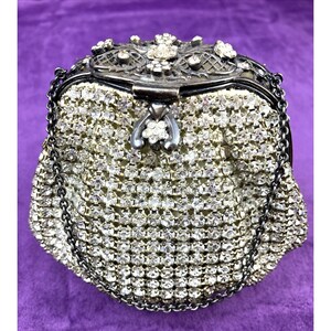 May include: A vintage, silver-toned evening bag encrusted with sparkling rhinestones. The bag features a decorative clasp and a delicate chain strap. The top frame is embellished with floral accents, adding to its elegant design. The bag's exterior is covered in a dense pattern of clear, round rhinestones.