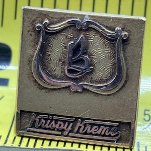 May include: A gold-colored metal stamp with a stylized butterfly design and the text "Krispy Kreme" in a cursive font.