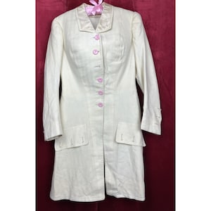 May include: Cream-colored long coat with a tailored design. Features a notched collar, long sleeves, and pink buttons down the front. Includes two front pockets and decorative details on the cuffs. The coat is hanging on a pink ribboned hanger.