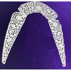 May include: A delicate, white lace collar with intricate floral and leaf patterns. The collar is composed of two main sections and a smaller piece, all connected. The lace is set against a deep purple background, highlighting the detailed design.