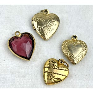 May include: Four heart-shaped pendants. One pendant features a faceted, deep red gemstone in a gold setting. The other three are gold-toned, with intricate designs, including floral and engraved patterns. Each has a loop for attaching to a chain.