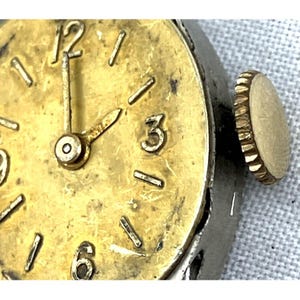 Op de afbeelding: Close-up van een vintage polshorloge met een goudkleurige wijzerplaat en wijzers. Het horloge heeft een zilverkleurige kast en een goudkleurige kroon. De wijzerplaat toont de cijfers 1, 3, 6, 9 en 12.