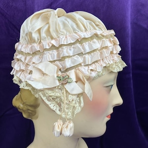 May include: A vintage cream-colored bonnet with layers of ruffled trim and lace edging. The bonnet features pale pink ribbon accents, a small floral embellishment, and two dangling satin pouches. The hat is displayed on a mannequin head.
