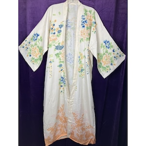 May include: A long, cream-colored robe featuring an open front and wide sleeves. The garment is decorated with a floral pattern in shades of blue, yellow, and green, with a soft orange leaf-like print along the bottom hemline.