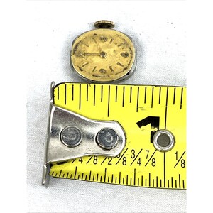 Op de afbeelding: Een klein, vintage polshorloge met een goudkleurige wijzerplaat en wijzers, tegen een witte achtergrond. Het horloge ligt naast een gele meetlint, met metingen in centimeters. De wijzerplaat van het horloge heeft zwarte markeringen.