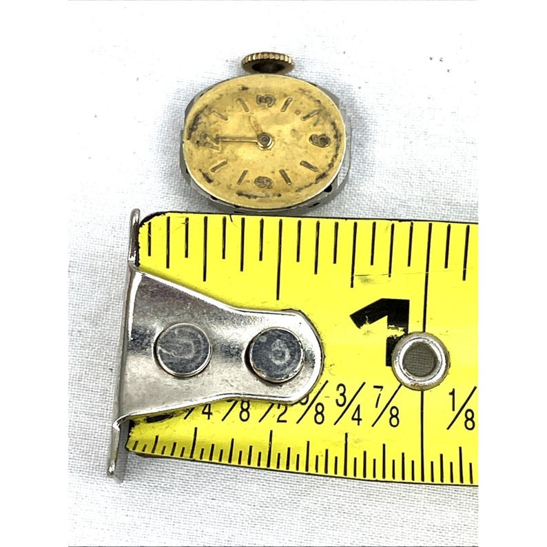 Op de afbeelding: Een klein, vintage polshorloge met een goudkleurige wijzerplaat en wijzers, tegen een witte achtergrond. Het horloge ligt naast een gele meetlint, met metingen in centimeters. De wijzerplaat van het horloge heeft zwarte markeringen.