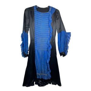 May include: A black long-sleeved dress featuring a central blue panel with horizontal smocking and vertical pleats. The dress includes blue ruffled trim along the side of the panel and blue ruffled cuffs on the sleeves, with a round neckline.