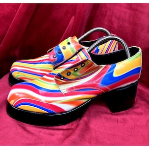 May include: A pair of colorful platform shoes with a rainbow striped pattern. The shoes have a black platform sole and a chunky heel. The uppers feature a vibrant mix of red, orange, yellow, green, blue, and white stripes. The shoes are a retro style.