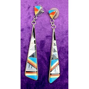 May include: A pair of silver-toned dangle earrings with a geometric design. Each earring features a triangular top with colorful inlays and a long, inverted triangle pendant with black, white, and turquoise accents. The earrings are displayed against a purple background.