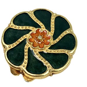 May include: A gold-toned, circular brooch with a dark green enamel design. The brooch features a central orange flower with a small crystal. The outer edge is detailed with small gold beads.